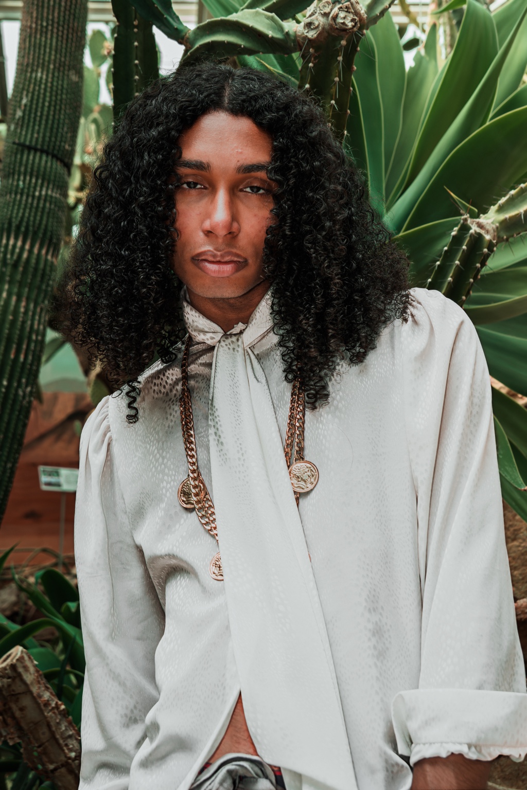 Jérémy Mounissamy, cheveux longs bouclés, chemise blanche et bijoux dorés, dans un jardin de cactus